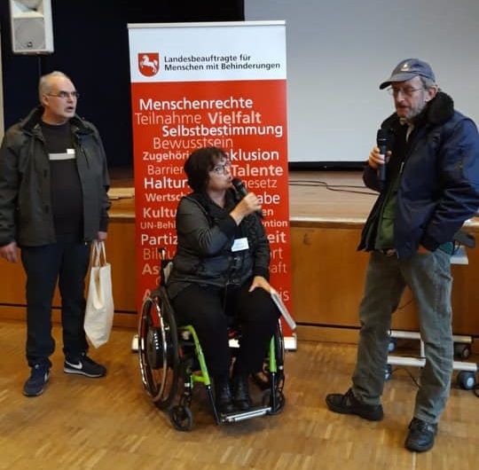 Vor der Bühne stehen Herr Kosciol, Frau Wontorra (im Rollstuhl) sowie Herr Altmann mit Mikrofonen in der Hand und begrüßen die Anwesenden.  Vor der Bühne stehen Herr Kosciol, Frau Wontorra (im Rollstuhl) sowie Herr Altmann mit Mikrofonen in der Hand und begrüßen die Anwesenden.